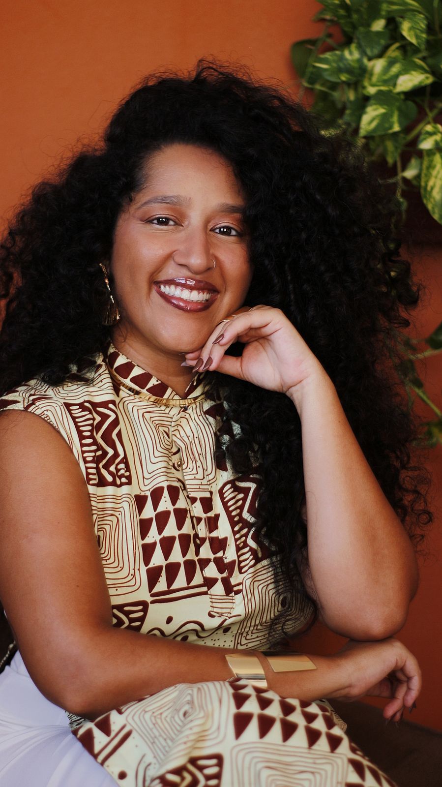 Retrato de Marina Niambi sorrindo, com vestido de estampa ancestral e parede terracota ao fundo